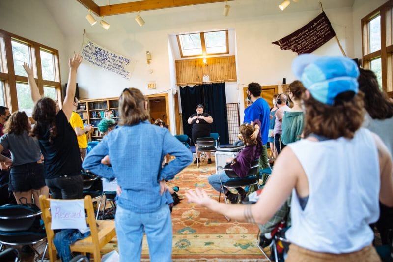 Jules, a fat white queer wearing all black, stands in a room with others who are standing with outstretched arms, they have both hands on their chest, teaching “Singing with Our Whole Selves” at Let My People Sing, Summer 2023